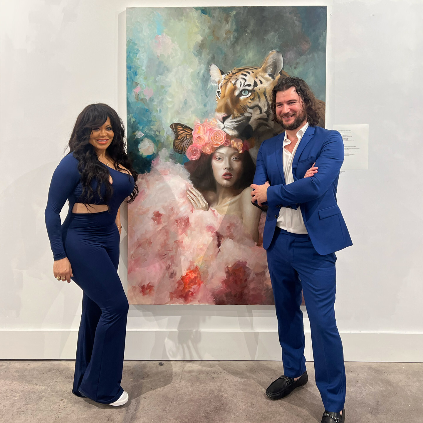 Artist Alex Righetto in a blue suit and Stacy in a blue outfit standing beside the 'The Guardian' painting at an art gallery
