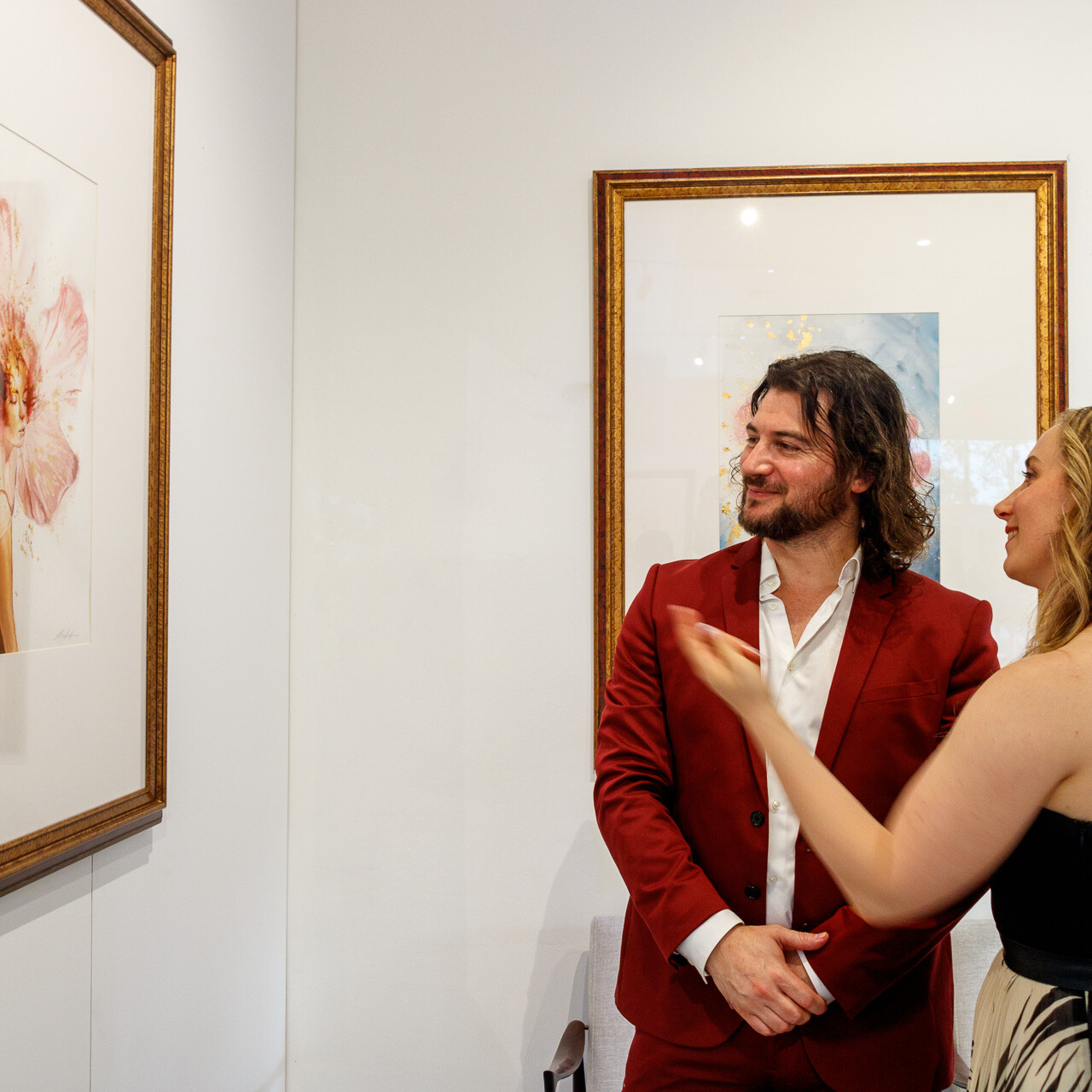 Artist Alex Righetto in a red suit discussing his 'Angelic Cherubic' painting from the Radiance Collection with a female art collector in a gallery.