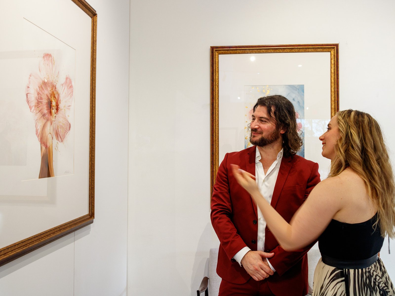 The Importance of Arts in our lives 12 Artist Alex Righetto in a red suit discussing his 'Angelic Cherubic' painting from the Radiance Collection with a female art collector in a gallery.