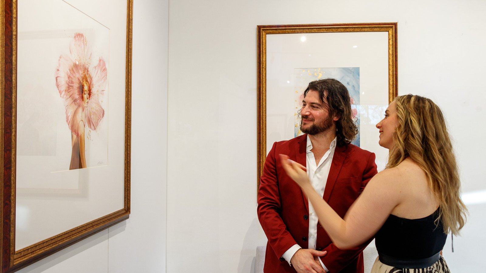 Artist Alex Righetto in a red suit discussing his 'Angelic Cherubic' painting from the Radiance Collection with a female art collector in a gallery.