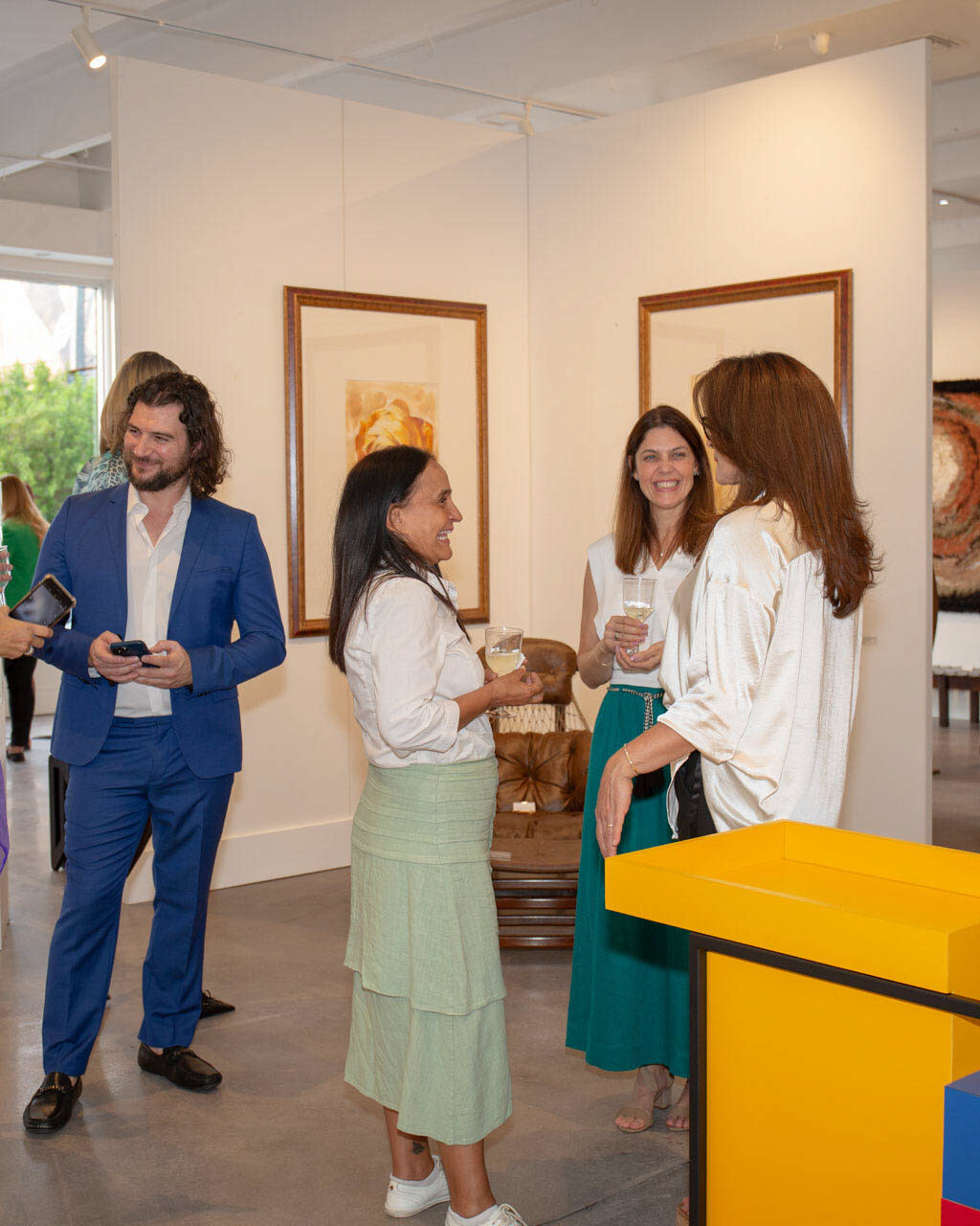 Artist Alex in a blue suit engaged in lively discussion with art collectors at a gallery exhibition, with paintings and sculptures in the background.