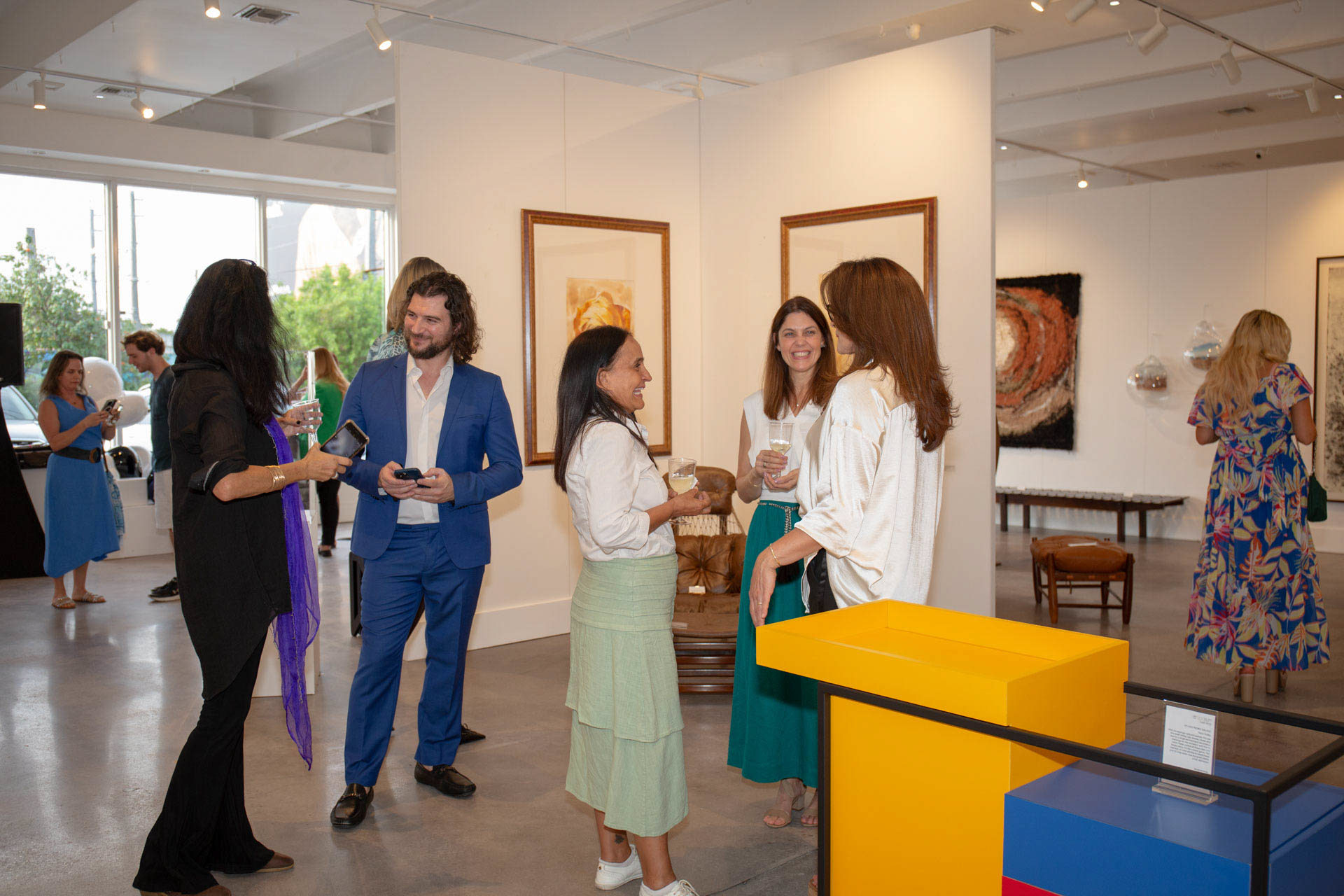 Artist Alex engaged in a lively discussion with art collectors at a gallery exhibition, with paintings and sculptures in the background.