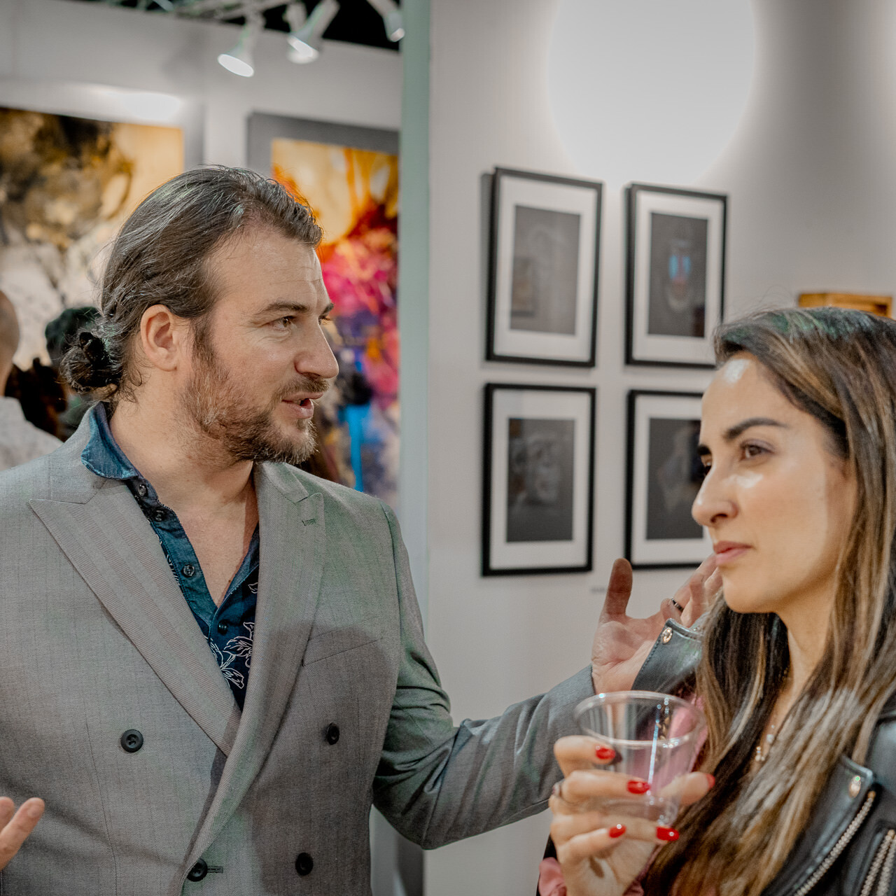 Artist Alex Righetto is seen engaging in a deep conversation with a visitor at the Spectrum Miami art fair, with his artwork in the backdrop.