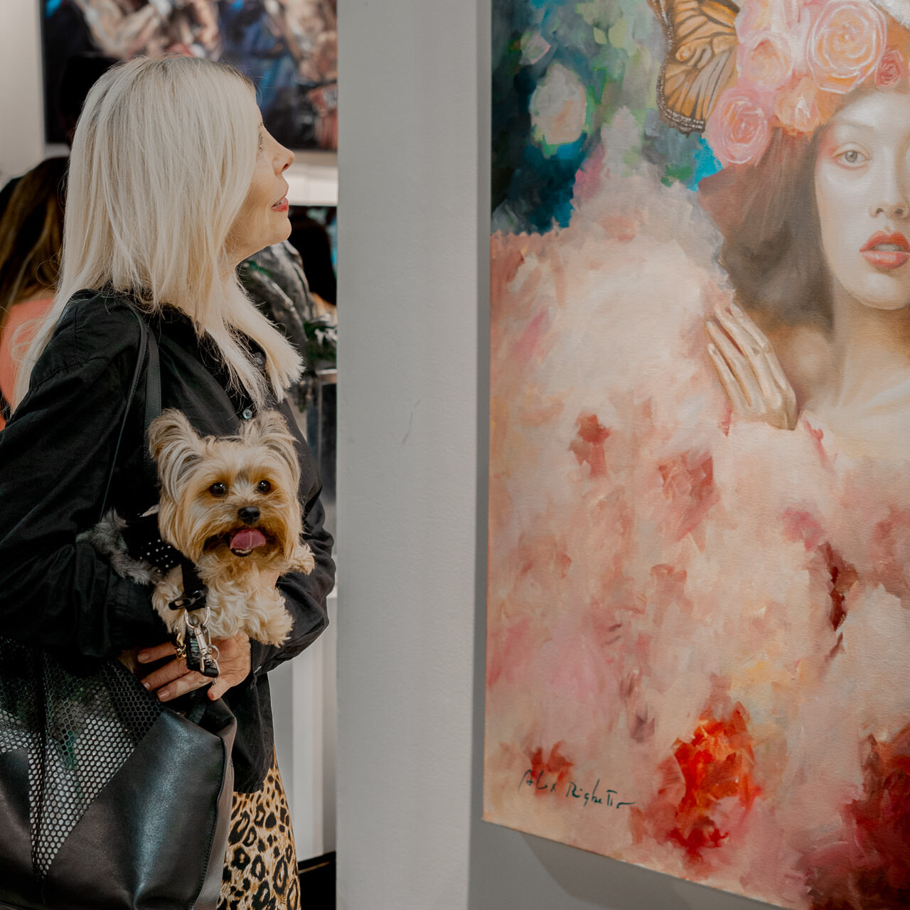 A woman with a small dog in her arms admires Alex Righetto's painting 'The Guardian' at an art exhibition, her attention absorbed by the artwork.