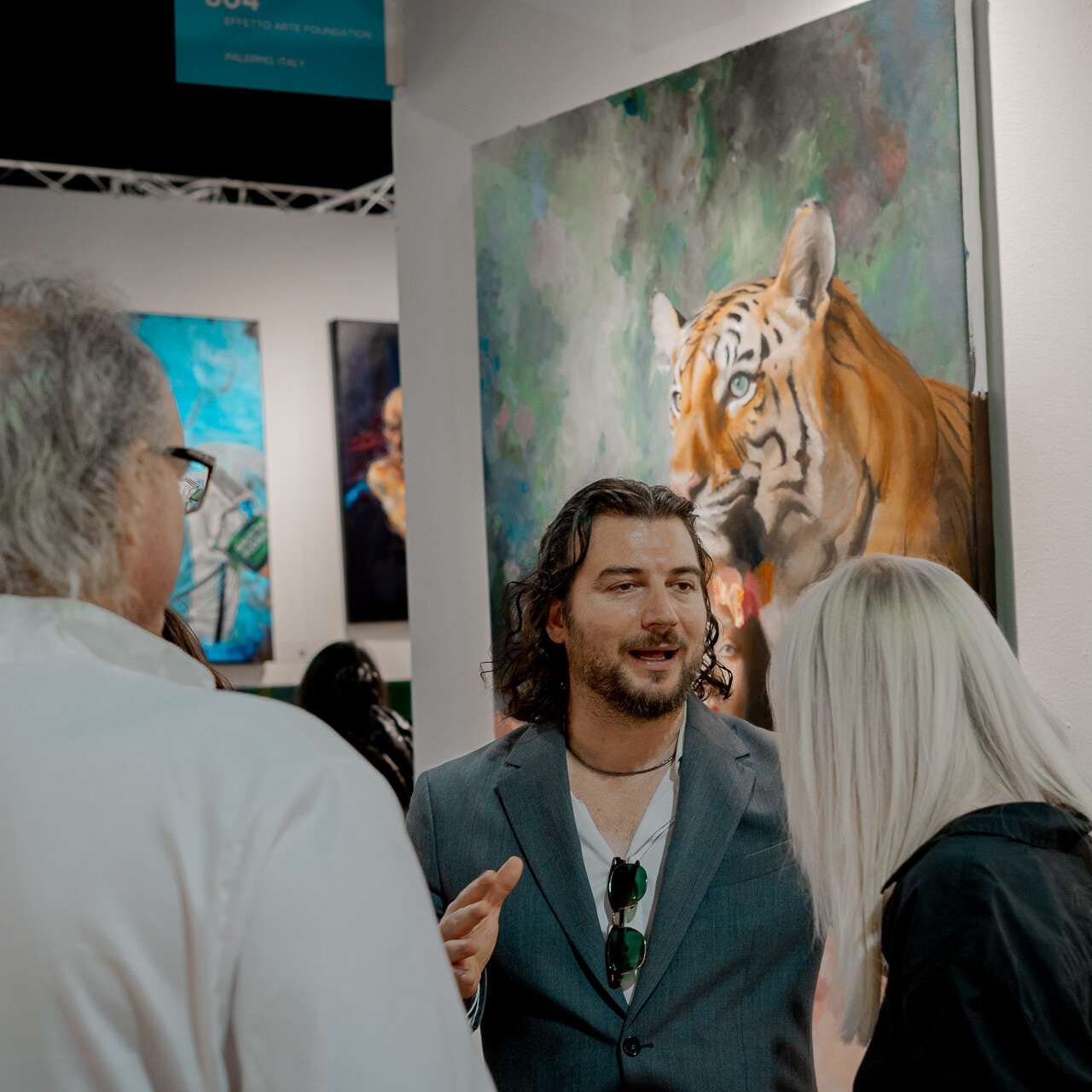 Alex Righetto is deeply engaged in conversation with visitors at Miami Art Week, with his expressive painting 'The Guardian' in the background.