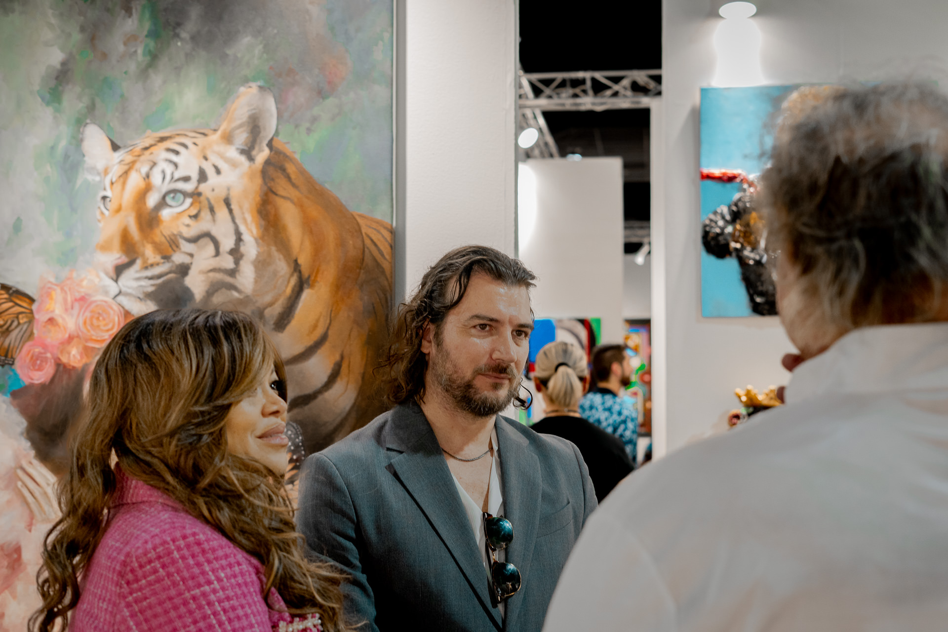 Artist Alex Righetto stands engaged with a group of attendees at Miami Art Week, with his painting 'The Guardian' adding a layer of artistic depth in the background.