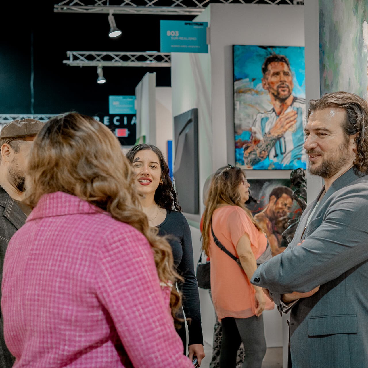 Artist Alex Righetto talks with visitors at an art exhibition, with his compelling painting 'The Guardian' displayed in the background.