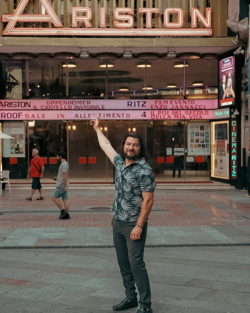 Alex Righetto smiling and pointing upwards in front of the Ariston Theatre's illuminated signboard at dusk