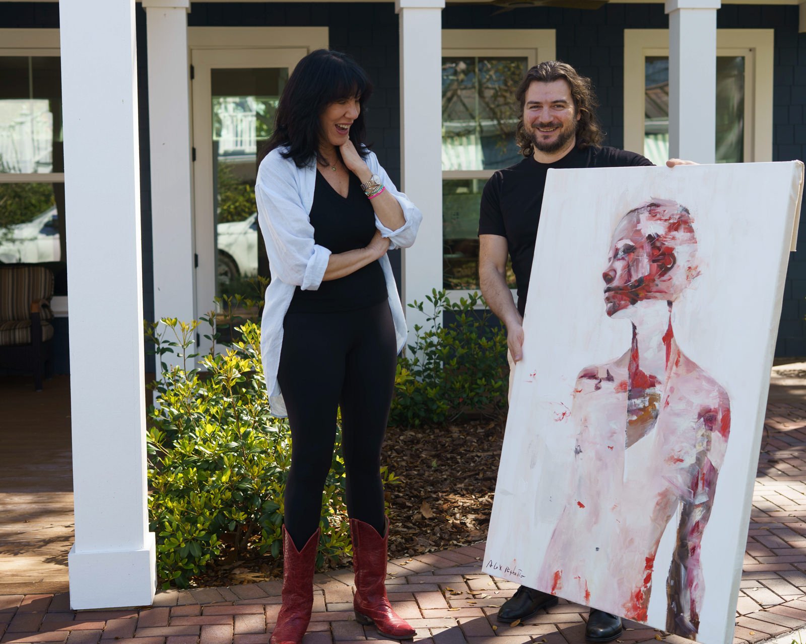 An artist smiles as he delivers an abstract painting to an enthusiastic collector. The woman, dressed in black with an open white shirt and red boots, laughs as she looks at the work. In the background is a house with a veranda and lush plants.  