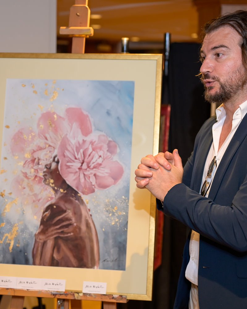 Alex Righetto Discussing the Radiance Collection 4 Close-up of Alex Righetto speaking with a collector, with a Radiance Collection watercolor artwork visible in the background at Art & Luxury Monaco 2026.